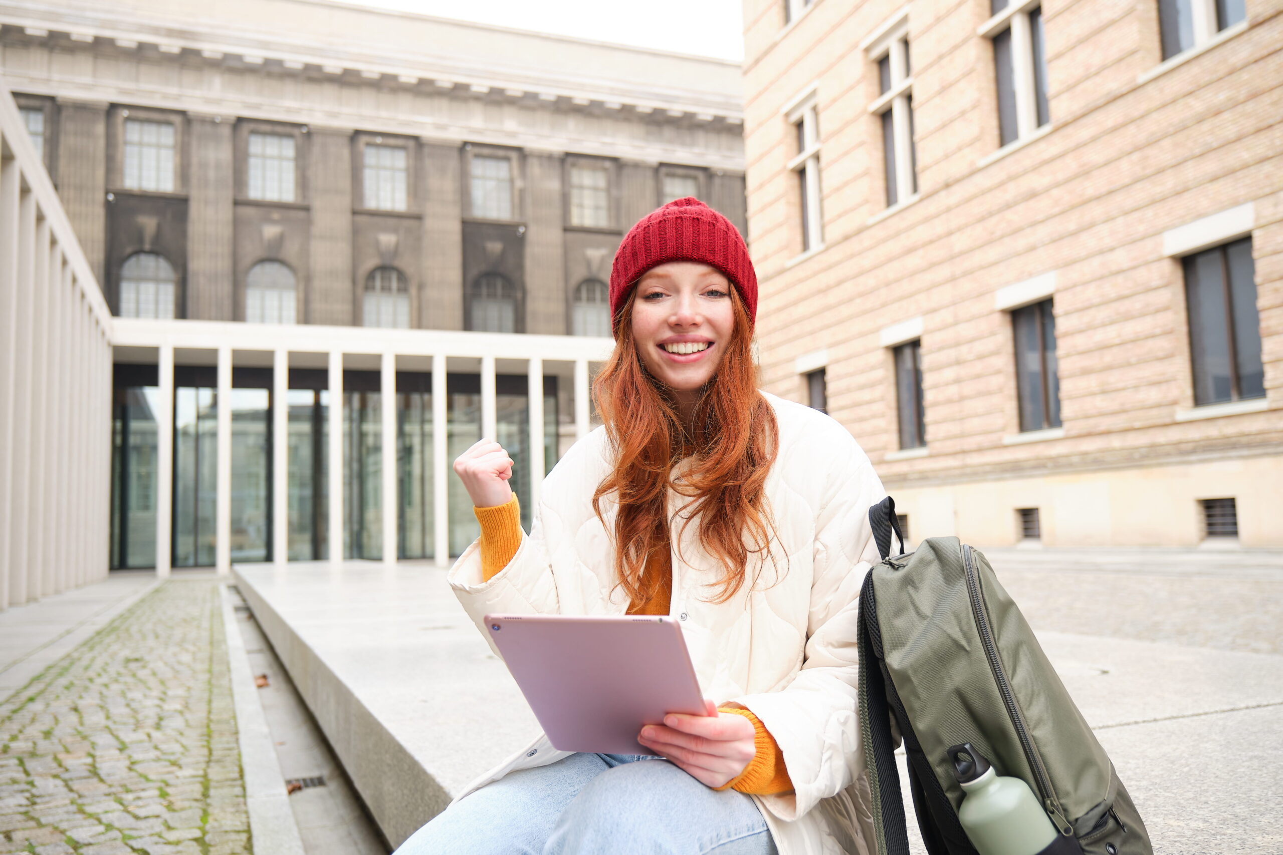beautiful-redhead-woman-red-hat-sits-with-backpack-thermos-using-digital-tablet-outdoors-conn